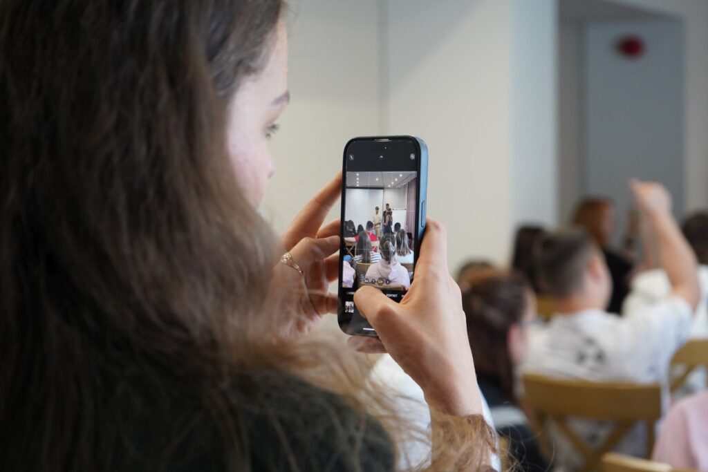 Uczestniczka wydarzenia fotografuje smartfonem spotkanie edukacyjne z udziałem młodzieży siedzącej na sali.